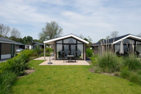 Photo of a modern lodge with glass front, patio seating, and lush garden on a partly cloudy day.