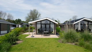 Photo d'un lodge moderne avec façade vitrée, terrasse aménagée et jardin verdoyant sous ciel nuageux.