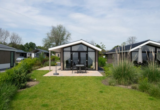 Photo of a modern lodge with glass front, patio seating, and lush garden on a partly cloudy day.