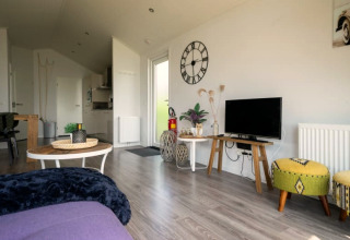 Helles Wohnzimmer einer Lodge mit TV, Wanduhr, grünen Hockern und moderner offener Küche im Hintergrund.