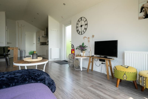 Modern lodge living room with TV, wall clock, green stools, wooden tables and open kitchen in the background.