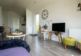 Modern lodge living room with TV, wall clock, green stools, wooden tables and open kitchen in the background.