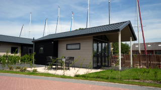 Modern lodge with patio, table, and chairs, situated near yachts with sailboat masts seen in the background.