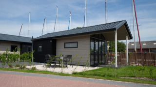 Moderne lodge med terrasse, bord og stole, placeret nær lystbåde med master synlige bag bygningen.