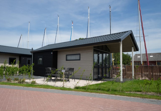 Lodge moderno con patio, tavolo e sedie, situato vicino a yacht con alberi velici visibili sullo sfondo.