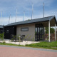 Moderne lodge med terrasse, bord og stole, placeret nær lystbåde med master synlige bag bygningen.