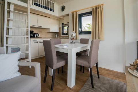 Interior de lodge moderno con mesa de comedor, cuatro sillas, cocina, escalera a altillo y ventana.