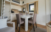 Moderne lodge met eettafel, vier stoelen, kitchenette, ladder naar vide, raam en beige gordijnen.
