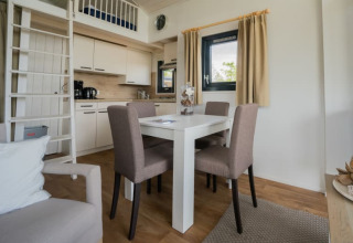 Moderne lodge met eettafel, vier stoelen, kitchenette, ladder naar vide, raam en beige gordijnen.
