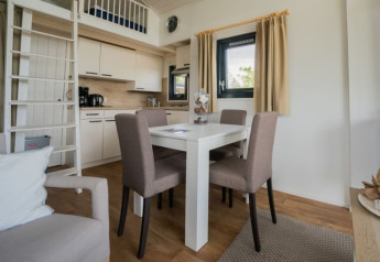 Moderne lodgeindretning med spisebord, fire stole, tekøkken og hems med stige, vindue og gardiner.