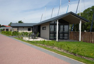 Et foto af moderne lodges med terrasse og udendørs siddeplads, omgivet af grønne planter og både i baggrunden.
