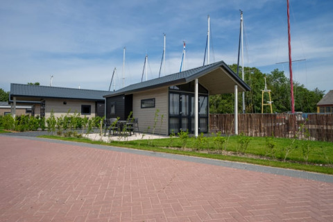 Een tiny house met overdekt terras en tuinmeubels, gelegen aan een weg, met zeilmasten op de achtergrond.