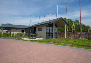 Et lille hus med overdækket terrasse og havemøbler, placeret ved en vej, med sejlmaster i baggrunden.