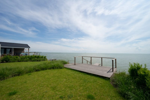 View of a lodge by the sea with a wooden deck, green lawn, and a wide sky filled with light clouds.