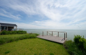 View of a lodge by the sea with a wooden deck, green lawn, and a wide sky filled with light clouds.
