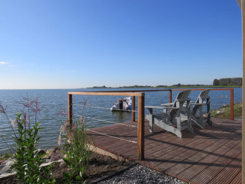 Wooden deck by the lake with two chairs and railing, overlooking water and a boat on a clear blue day.