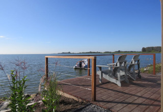 Wooden deck by the lake with two chairs and railing, overlooking water and a boat on a clear blue day.