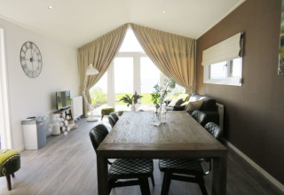 Modern lodge interior with a dining table, leather chairs, sofa corner and large windows facing greenery.