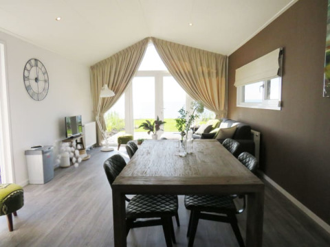 Intérieur de lodge moderne avec salle à manger, chaises en cuir, canapé et grandes fenêtres donnant sur la verdure.