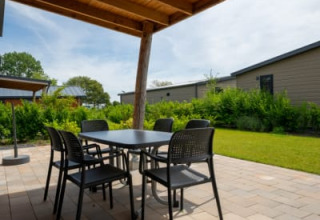 Patio coperto di una lodge con tavolo, sedie e pavimento in pietra, circondato da prato e vegetazione.