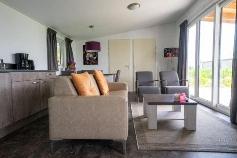 Moderne lodge-stue med sofa, lænestole, sofabord og store vinduer, der giver udsigt til grønne omgivelser.