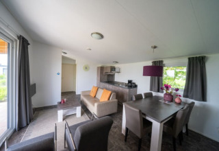 Interno lodge luminoso e moderno con divano, tavolo da pranzo, cucina e ampie finestre con tende grigie.