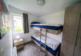 Kleine lodge slaapkamer met stapelbed, nachtkastje, raam met gordijn, kast en muurschilderij aanwezig.