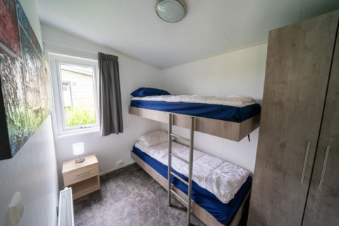 Dormitorio pequeño en una cabaña con litera, mesa de noche, ventana con cortina, armario y cuadro en la pared.