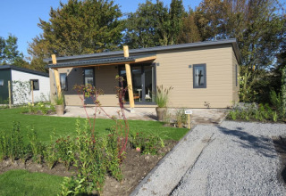 Moderne lodge med træbeklædning og overdækket terrasse, omgivet af grønt og nydeligt anlagt have.