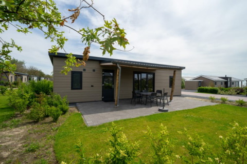 Acogedora cabaña con terraza y muebles de exterior, rodeada de vegetación en un parque vacacional tranquilo.