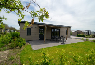 Acogedora cabaña con terraza y muebles de exterior, rodeada de vegetación en un parque vacacional tranquilo.