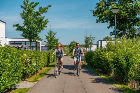 Twee mensen fietsen op een zonnige dag door een vakantiepark met groene hagen en moderne glamping huisjes.