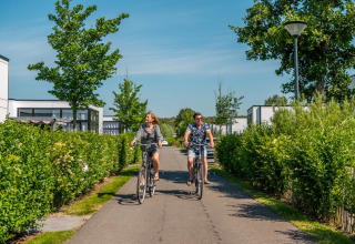 Twee mensen fietsen op een zonnige dag door een vakantiepark met groene hagen en moderne glamping huisjes.