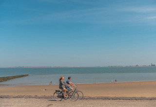 Twee mensen fietsen langs het strand bij een vakantiepark met glamping accommodaties onder een blauwe lucht.
