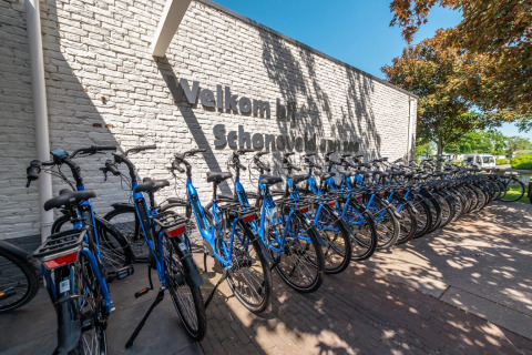 Rij blauwe fietsen geparkeerd bij een witte bakstenen muur met een welkomstbord bij een vakantiepark.