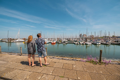 Twee mensen kijken uit over een jachthaven vol boten bij een vakantiepark met glamping accommodaties.