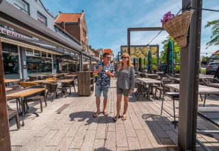 Twee mensen wandelen met ijsjes op een zonnig terras bij een vakantiepark met glamping accommodaties.