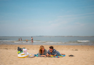 Twee mensen ontspannen op een strand in een vakantiepark met glamping, met opblaasbare speelgoed in de buurt.