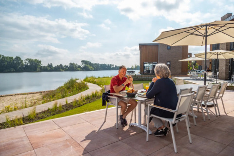 Twee mensen genieten van een maaltijd op een terras aan het water in een vakantiepark met glamping.
