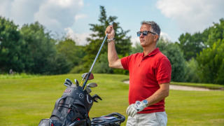Man in rode polo op een golfbaan, haalt een golfclub uit tas, omringd door groen en zonnig weer.