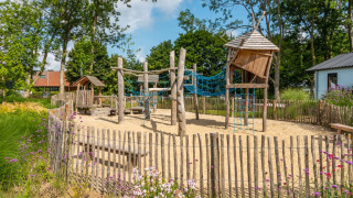 Buiten speeltuin met houten speeltoestellen en zand omringd door een houten hek bij een vakantiepark.