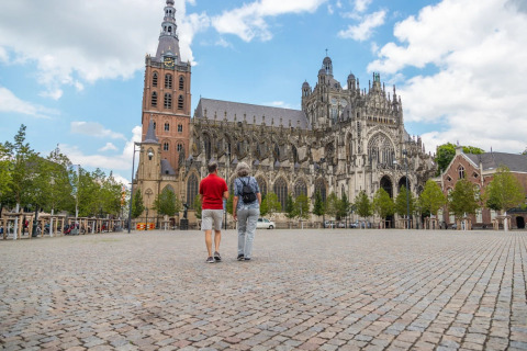 Twee mensen wandelen over een plein met zicht op een grote gotische kathedraal en toren, omgeven door bomen.
