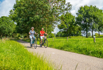 Twee mensen fietsen samen op een rustig pad door een vakantiepark met veel groen en bomen, zonnige dag.