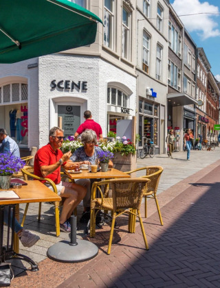 Mensen genieten van een zonnig terras in een winkelstraat met bloemen, fietsen en gezellige sfeer.