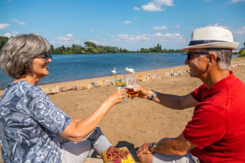 Twee oudere mensen genieten van drankjes aan het strand van een vakantiepark met glamping, bij een meer.