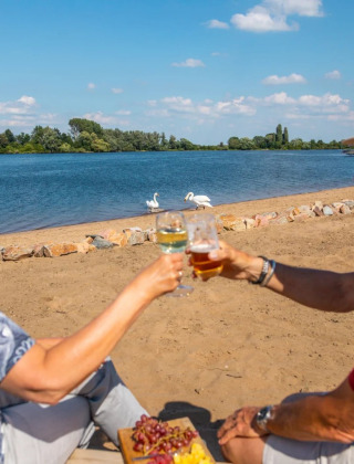 Twee oudere mensen genieten van drankjes aan het strand van een vakantiepark met glamping, bij een meer.