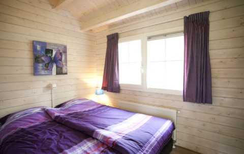 Acogedor dormitorio de cabaña con paredes de madera, cama doble con ropa de cama morada y ventana grande.