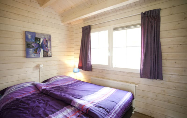 Acogedor dormitorio de cabaña con paredes de madera, cama doble con ropa de cama morada y ventana grande.