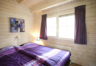 Acogedor dormitorio de cabaña con paredes de madera, cama doble con ropa de cama morada y ventana grande.