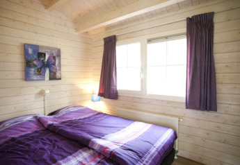 Chambre confortable dans un lodge, murs en bois, lit double violet, grande fenêtre et rideaux violets.